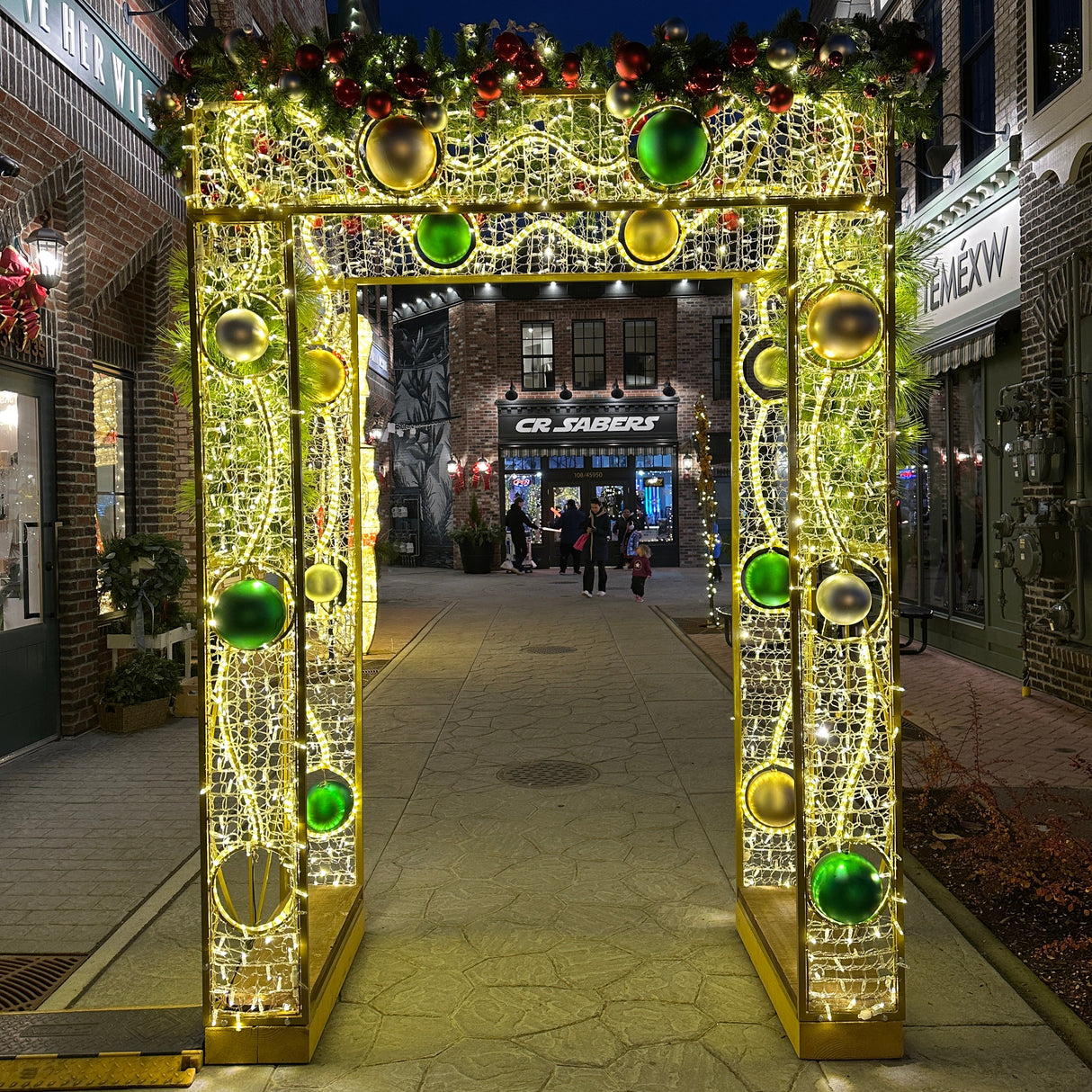 A decorative archway with twinkling lights and green ornamental elements, leading to a bricked pathway with a festive display.